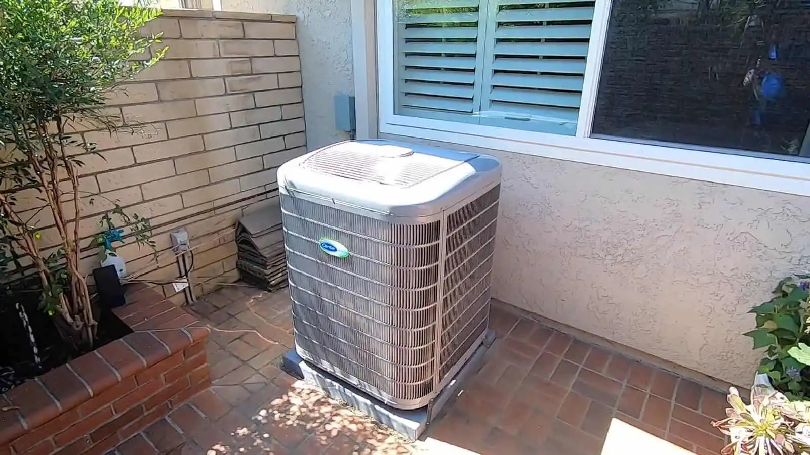 AC unit on a brick patio outside a house with a window and plants.